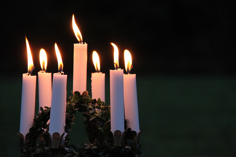 Seven white candles are burning in a circular arrangement with green foliage around the base, set against a dark background