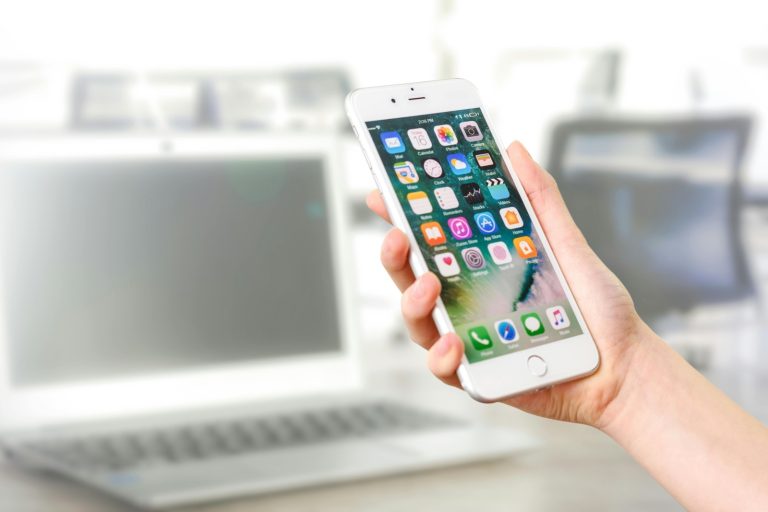 A person holds a smartphone with the home screen visible, displaying various app icons
