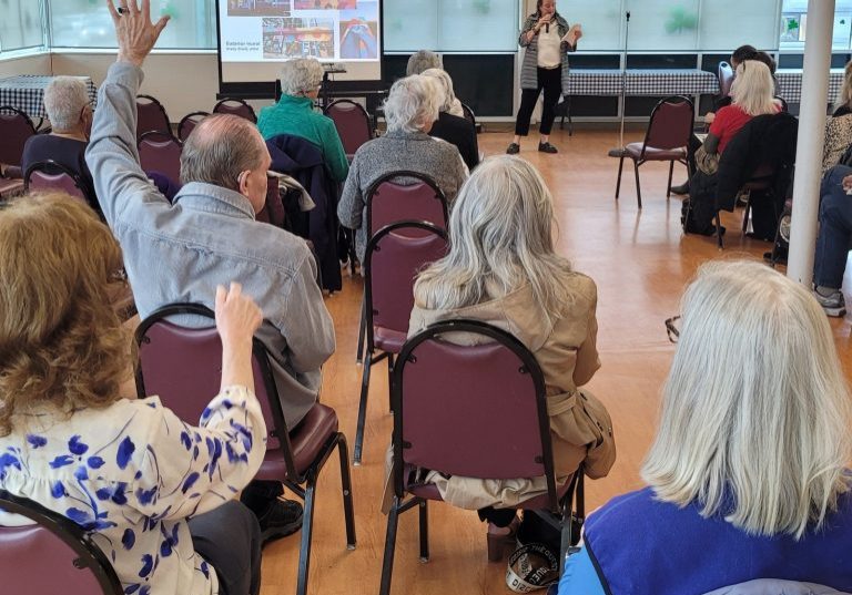 In a membership meeting, a group of people seated in a room listens intently to the speaker near the presentation screen. One person raises a hand, likely for a question or to ask for a quick recap.