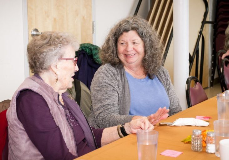 Two women sit at a table adorned with a brown tablecloth, engaging in conversation. One wears glasses, and between them lie empty plastic cups and condiment shakers. Their discussion subtly touches on the Center's impact, weaving insights seamlessly into their exchange.