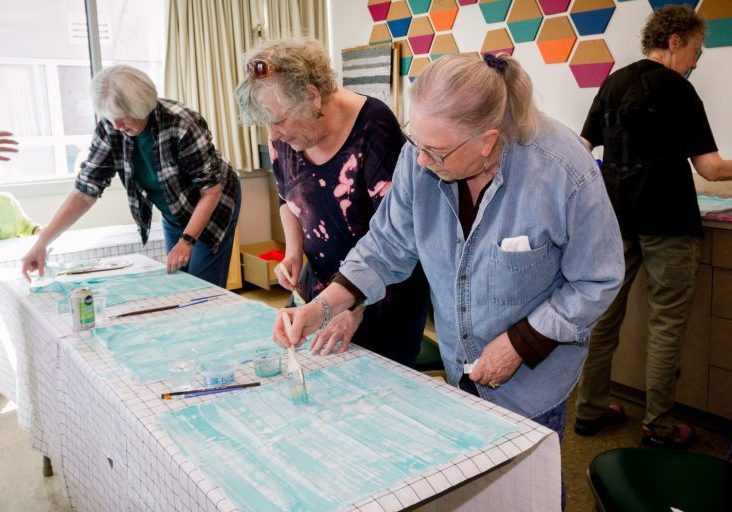 Photo from The Center's fabric painting class with several people standing and working on their projects