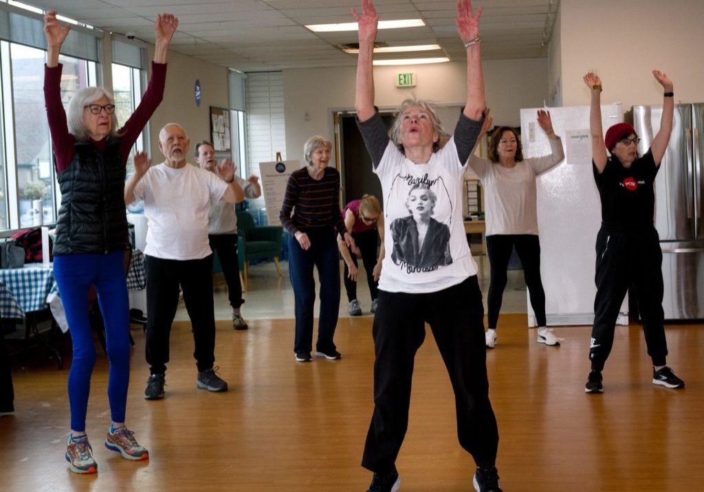 A group of adults participate in a wellness-focused fitness class, raising their arms while standing in Hatten Hall at the Center