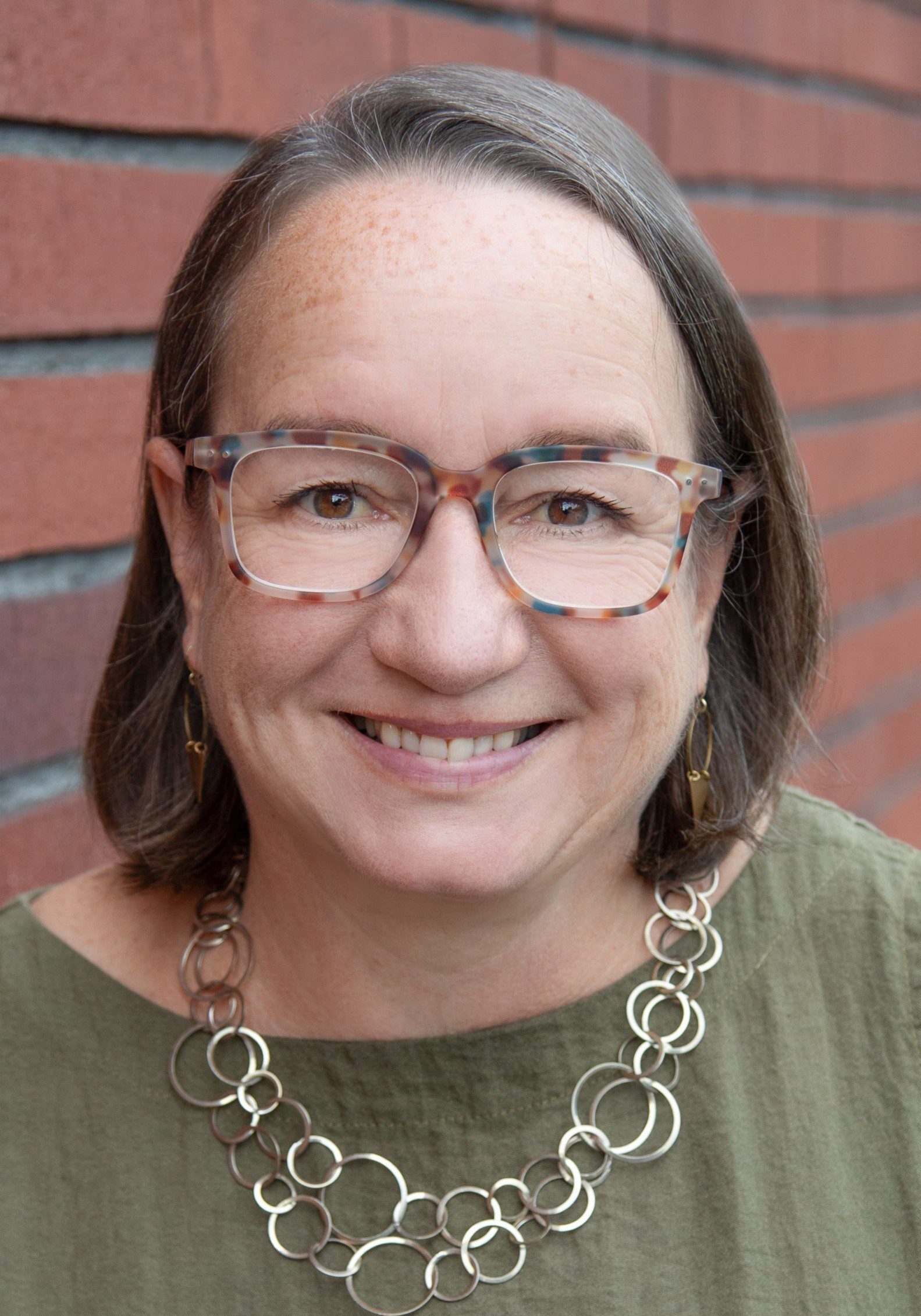 Headshot of Amy Lee, with glasses, shoulder-length brown hair, and a green sleeveless top smiles while standing against a red brick wall