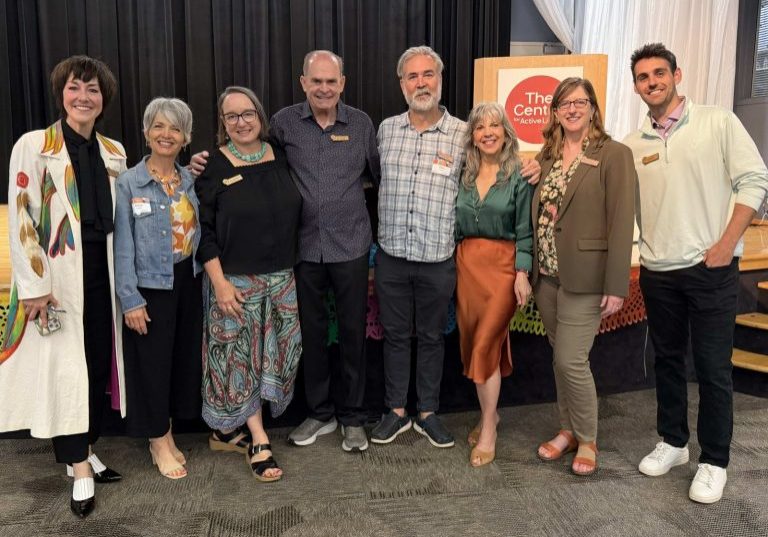 7 of The Center's board members stand side by side, smiling in front of a podium labeled at the Annual Fundraising Breakfast