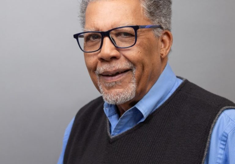 Headshot of Dennis Jackson, an older black man with gray hair and a goatee wearing glasses and a blue collared shirt with dark sweater vest.