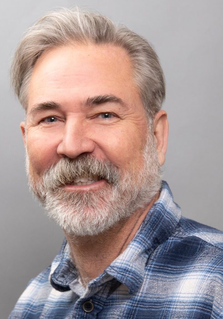 Scott Collins smiles at the camera. He is wearing a blue plaid shirt and stands against a plain gray background, embodying the friendly spirit of the Board at The Center.