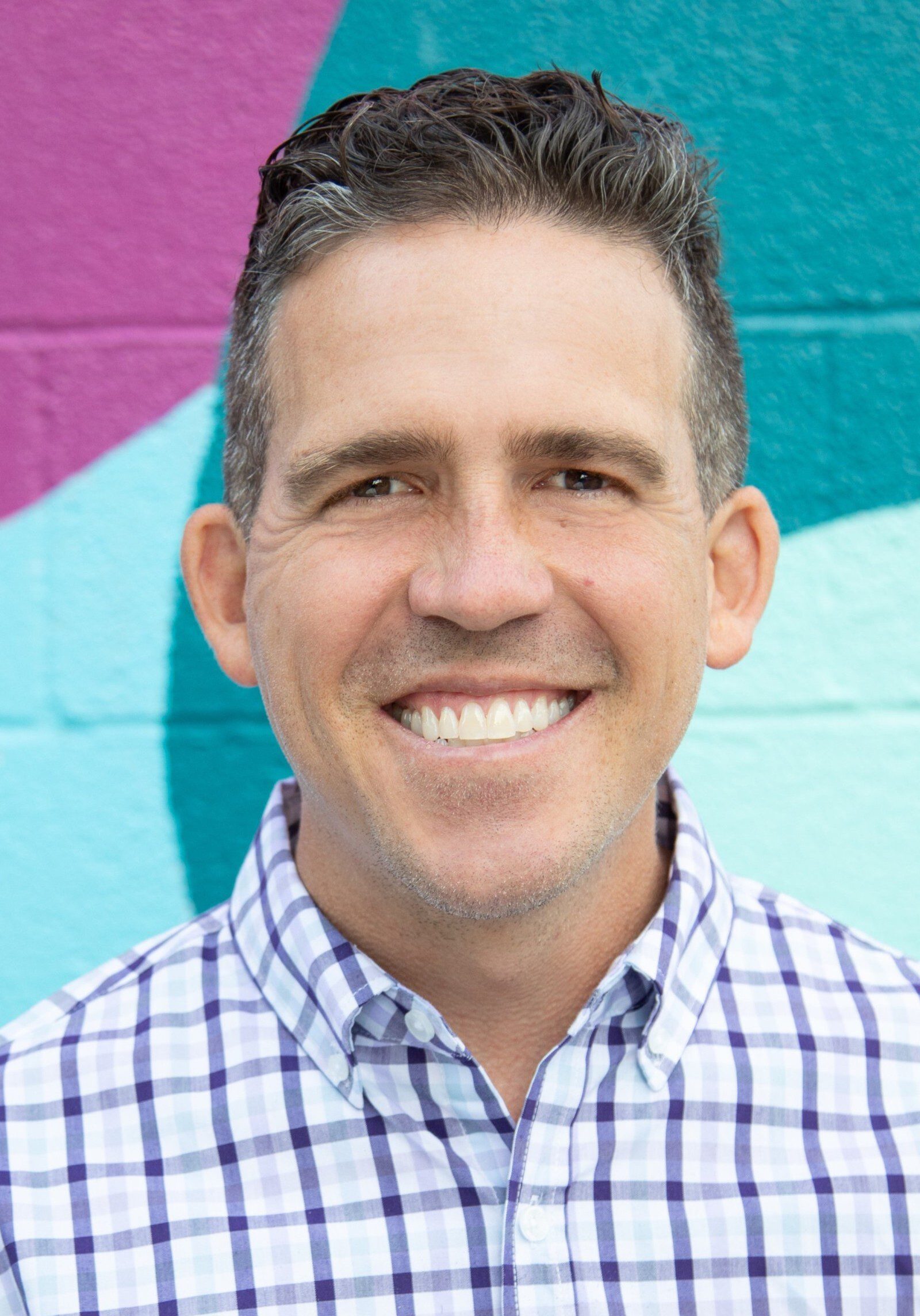 Christopher with short dark hair, a board staff member, smiles at the camera, wearing a plaid shirt, standing in front of a colorful mural with purple, teal, and light blue shapes.