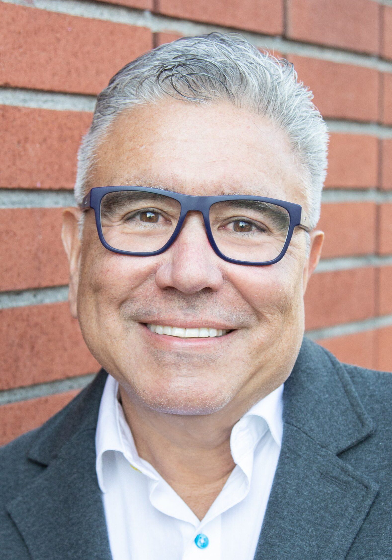 Danny with gray hair and glasses, dressed in a white shirt and dark blazer, smiles in front of a red brick wall—capturing the approachable spirit of our staff.