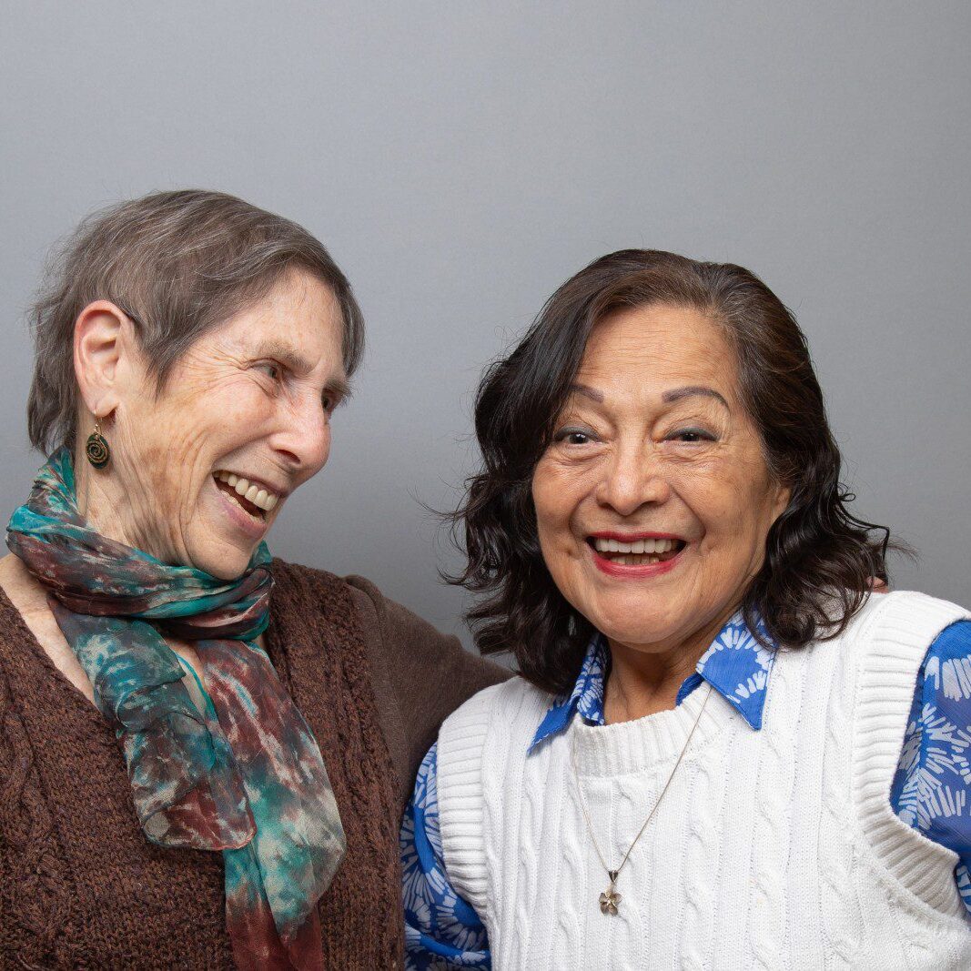 Two women stand close together, smiling and laughing in front of a plain gray background.