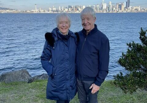 Volunteers Ceedee and Bob, both older adults in jackets, stand side by side on a grassy area by the water, with a city skyline visible in the background under a blue sky.