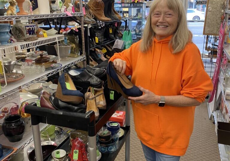 A Thrift store volunteer in an orange hoodie smiles while holding a shoe in a thrift store aisle filled with summer finds—various shoes, ceramics, and household items.