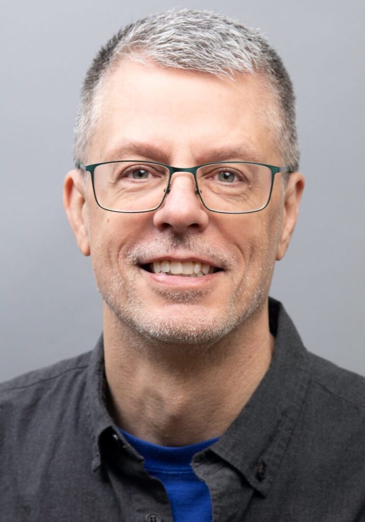 Jack Jannack smiles at the camera. He is wearing a black collared shirt over a blue shirt against a neutral grey background, fitting seamlessly among the dedicated Staff at The Center.