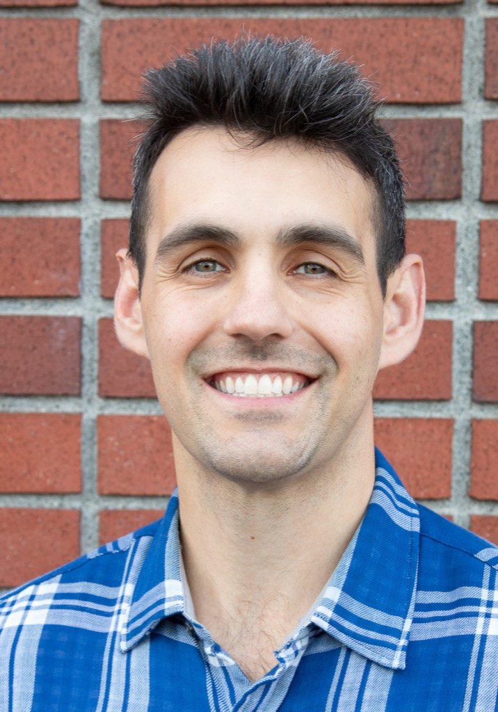Jack has short dark hair and a blue plaid shirt, who is part of the staff, smiles in front of a red brick wall.