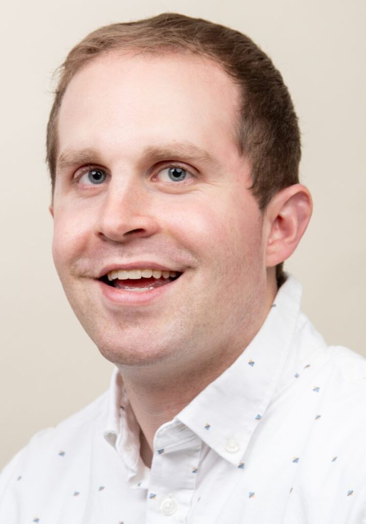 Josh Maduell, smiling, wearing a white button-up shirt with small blue patterns, poses against a neutral background. He is part of the Staff at The Center.