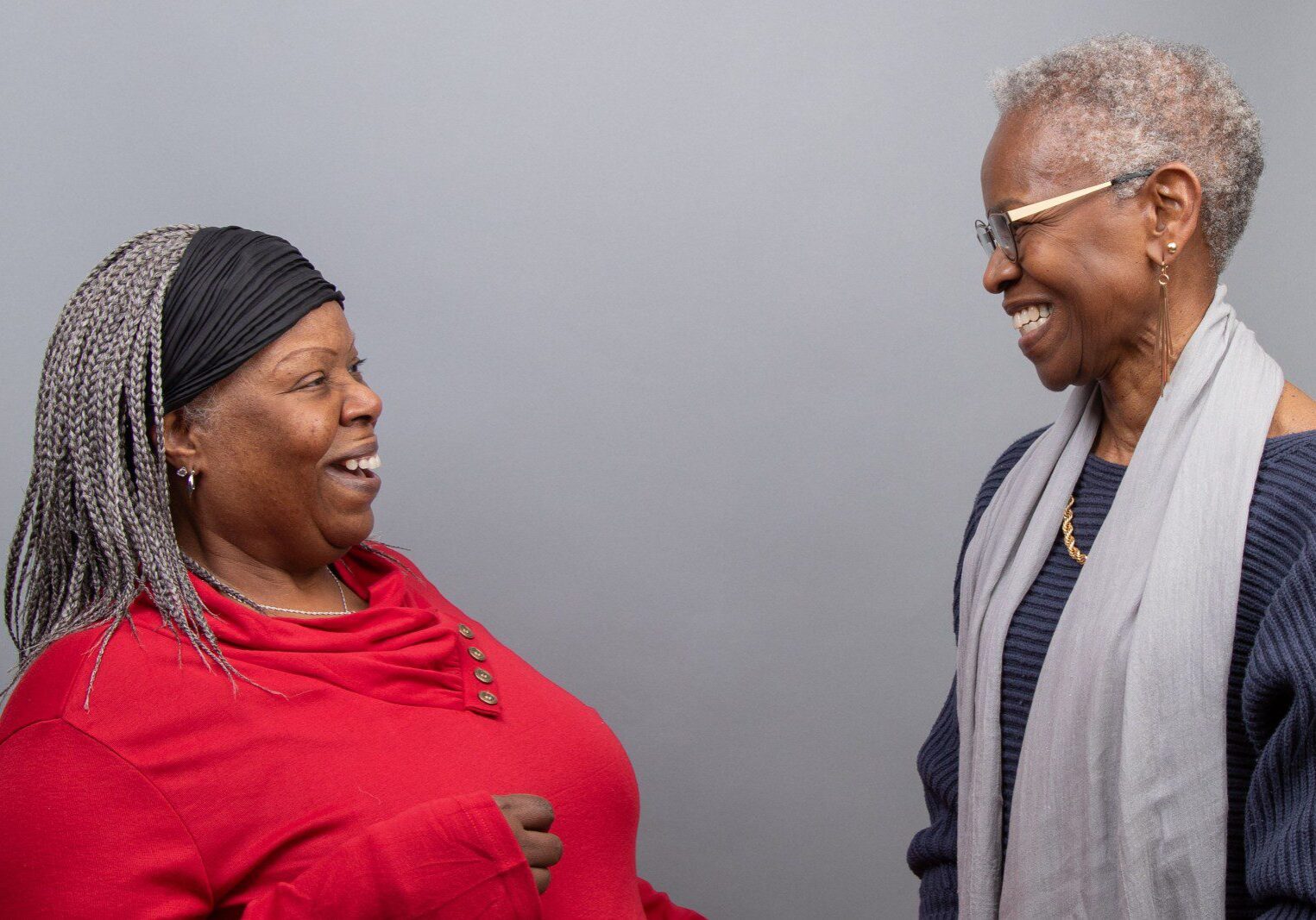 Two women stand facing each other, smiling and laughing, against a plain gray background.