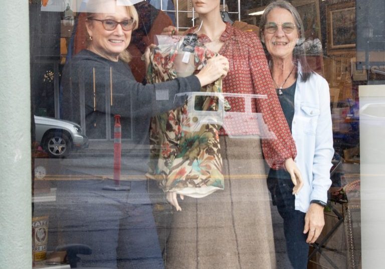 Two women are thoughtfully arranging a mannequin in the thrift store's window display.