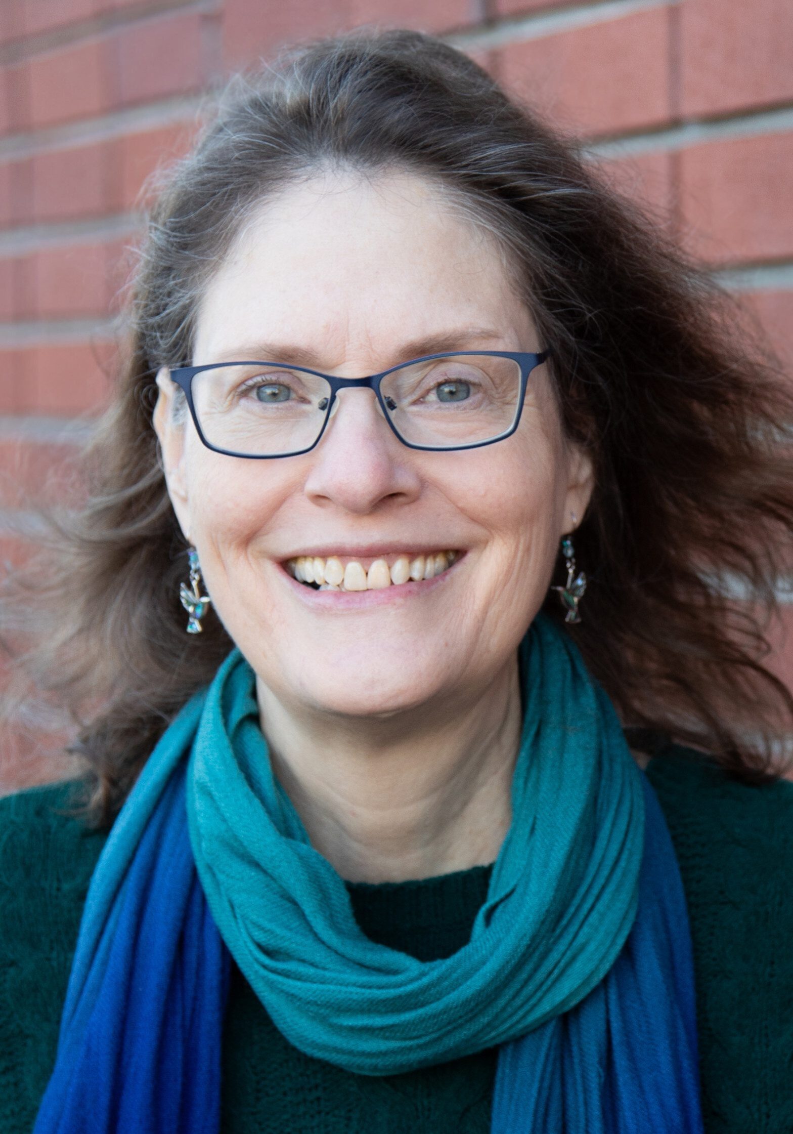 Michele Fawcett-Long, a member of the Staff at The Center, with glasses, long hair, and a blue scarf, smiles in front of a red brick wall.