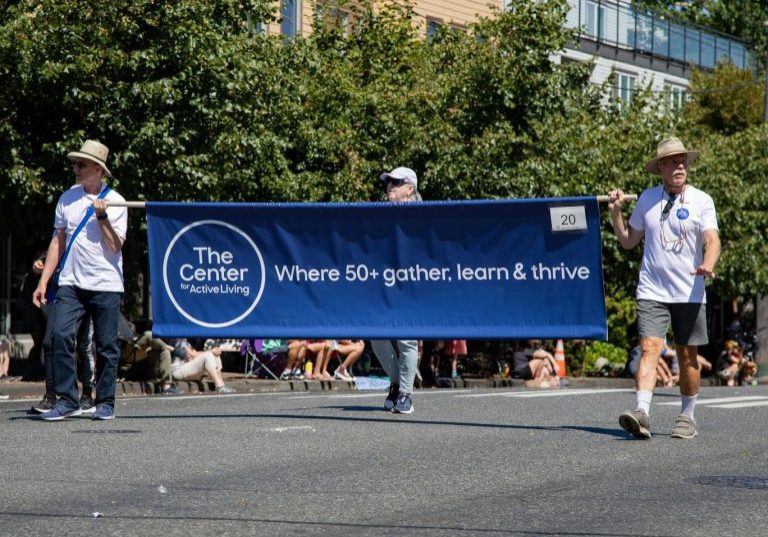Center members march in the West Seattle parade holding a blue banner that reads, "The Center for Active Living: Where 50+ gather, learn & thrive."