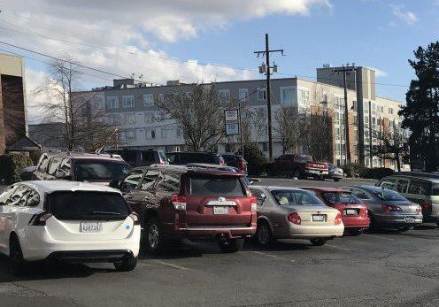 Several cars are parked in a pay parking lot on a sunny day, with an apartment building and trees visible in the background.