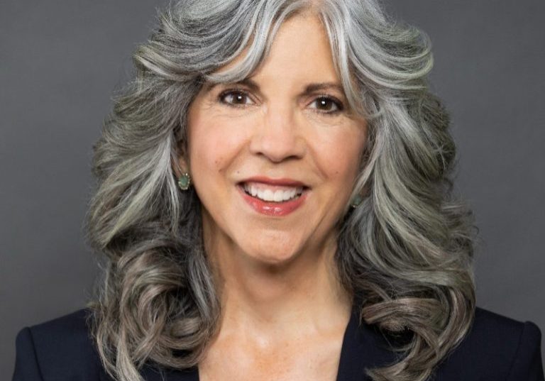 Steph Bruno with shoulder length, wavy gray hair and a black blazer smiles against a gray background