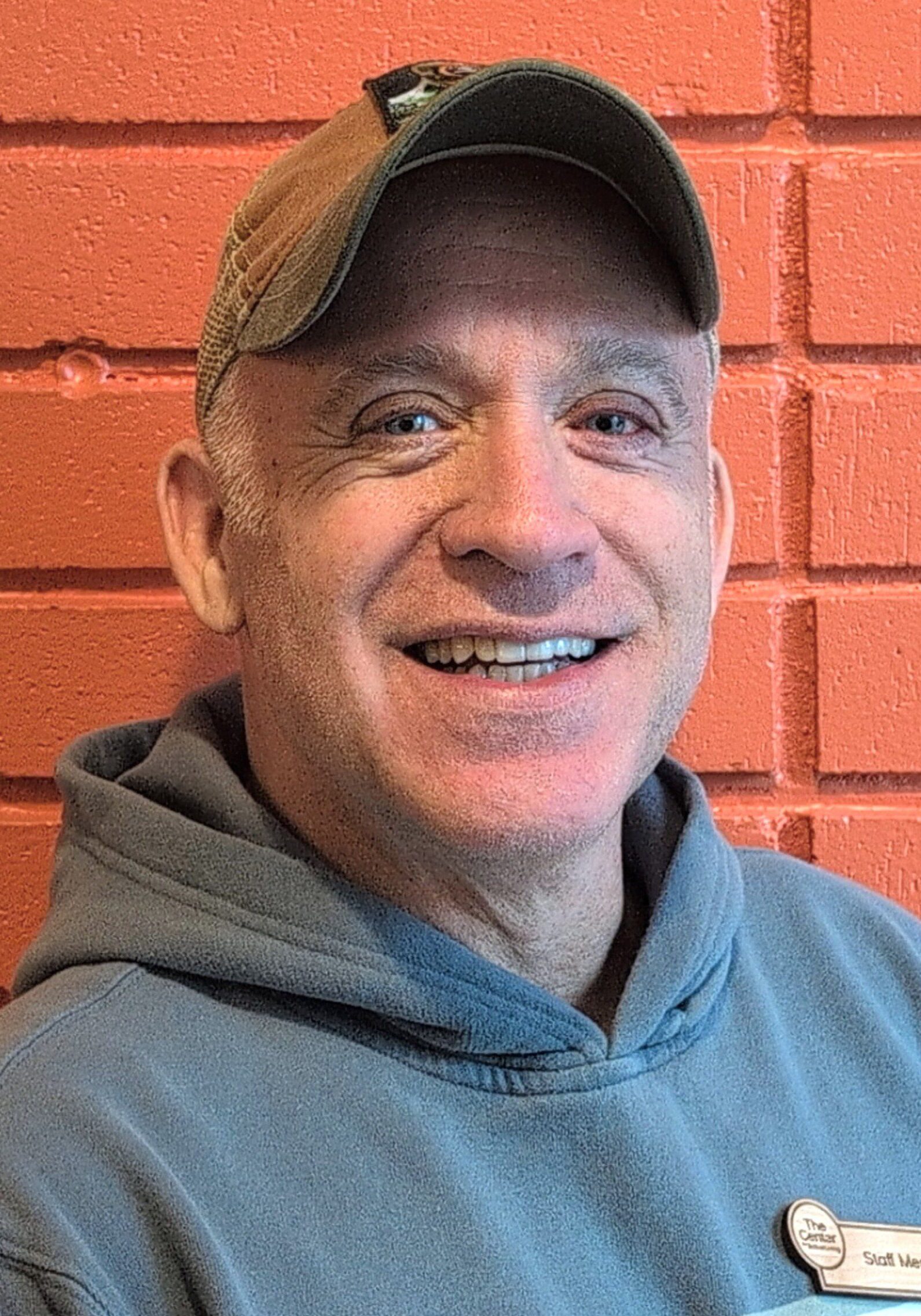 Terry wearing a baseball cap and hoodie smiles while sitting in front of a red brick wall, looking like a friendly staff member on break.