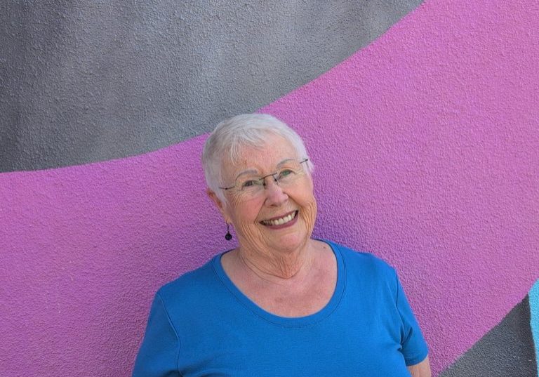 Susana Herbert with short white hair and glasses, smiles as she stands in front of a colorful wall with gray and pink curved shapes, wearing a blue shirt.