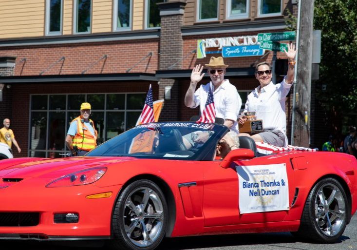 Neil and Bianca wave from a red convertible Corvette, proudly adorned with small American flags and a sign on the door. As volunteers in the parade, they cruise past a brick building, smiling at the cheering spectators in the background.