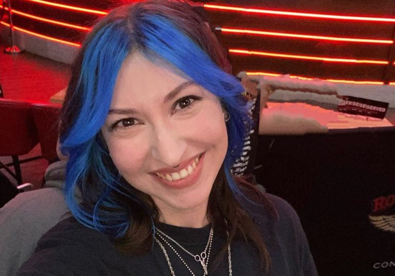 Photo of Yoni with blue-streaked hair smiles for a selfie in front of a stage with musical instruments and red lights in the background.