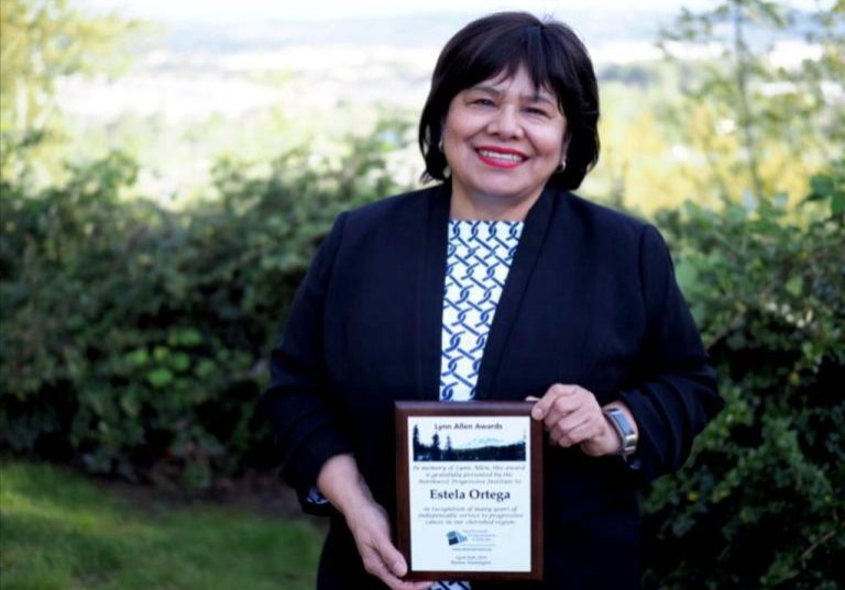 Estela Ortega in a dark blazer stands outdoors holding a framed award plaque and smiling at the camera