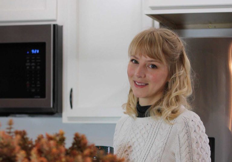 A graceful figure with blonde hair stands in the kitchen, wearing a white sweater. Nearby, a microwave and cabinets blend into the scene, while a vibrant plant adds life to the foreground. Perhaps she's not just cooking but volunteering her time for something greater.