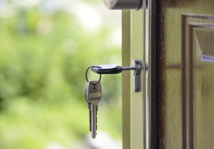 A set of keys is inserted in the lock of a slightly open wooden door, with greenery visible in the blurred background