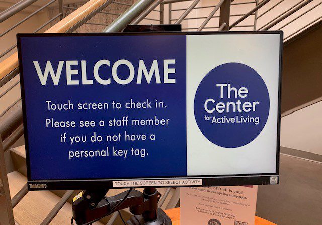 A computer screen displays a welcome message and center membership check-in instructions for The Center for Active Living, next to a staircase railing.