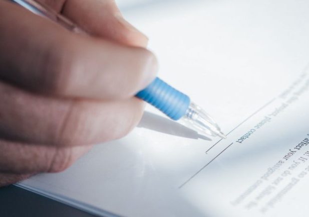 A hand holds a blue pen, signing a document on a white sheet of paper with printed text